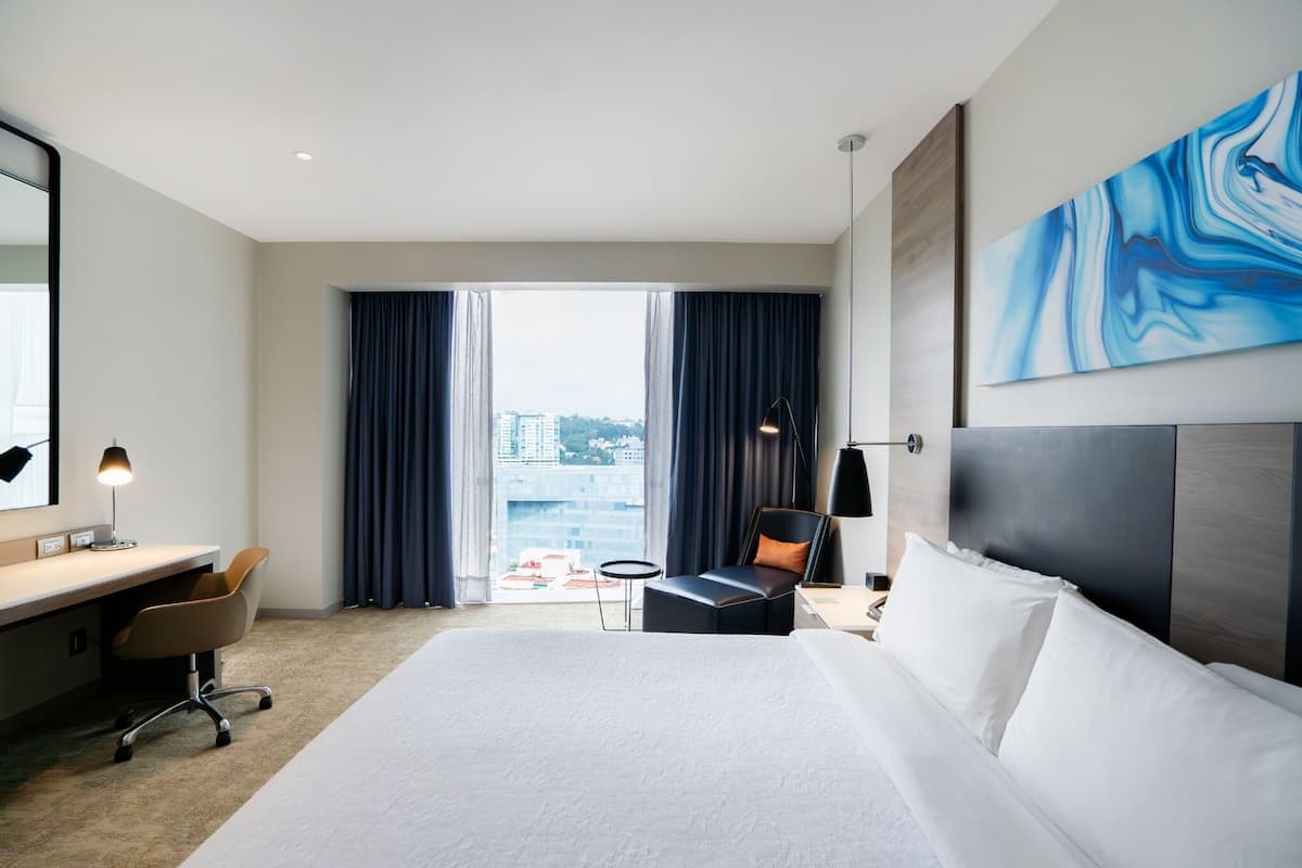 A hotel room with a large bed, a desk with a chair, a small table, a lamp, and a view of the city through large windows.