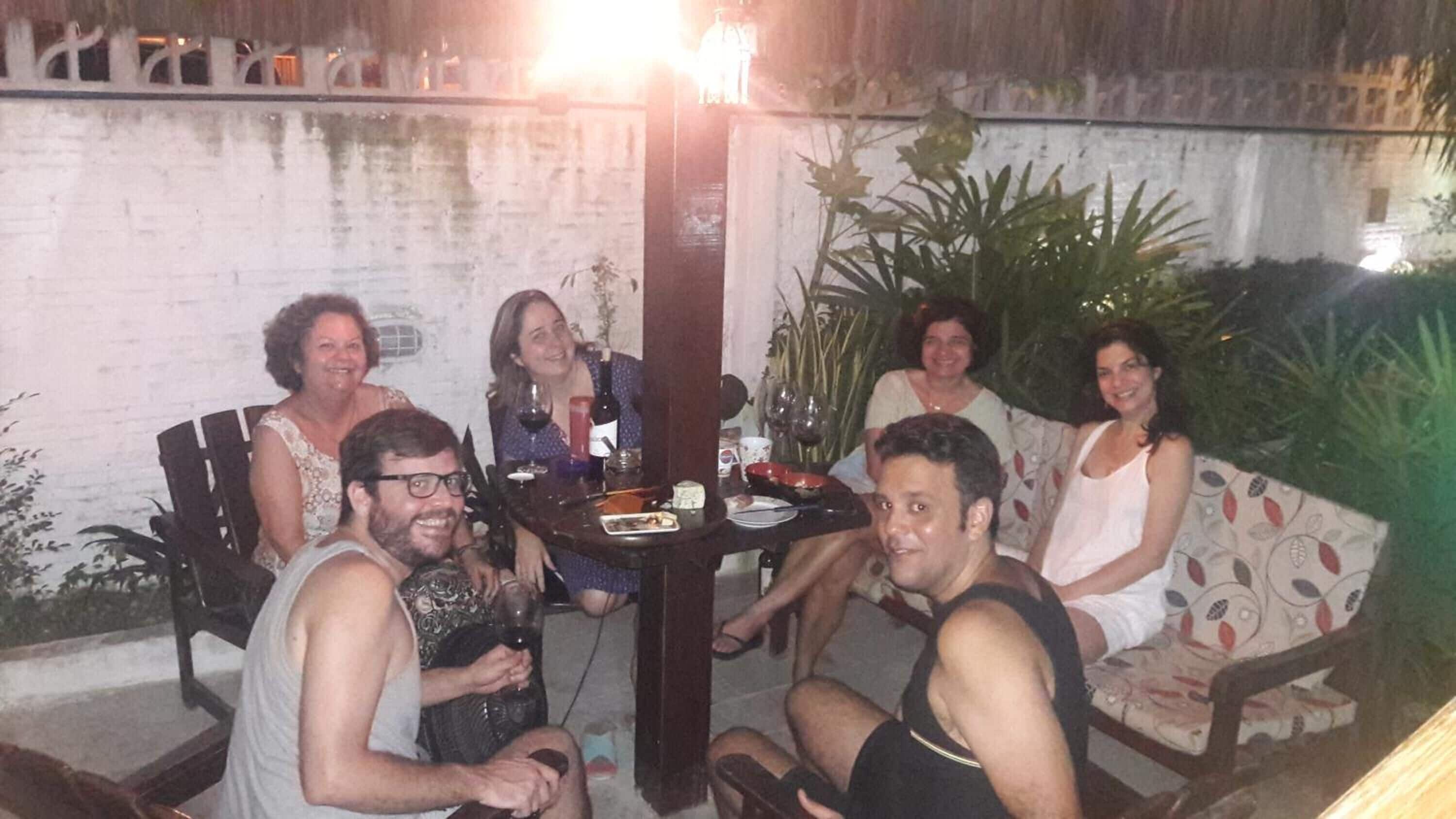 A group of people seated around a table with drinks and snacks, in an outdoor setting with a brick wall and plants in the background.