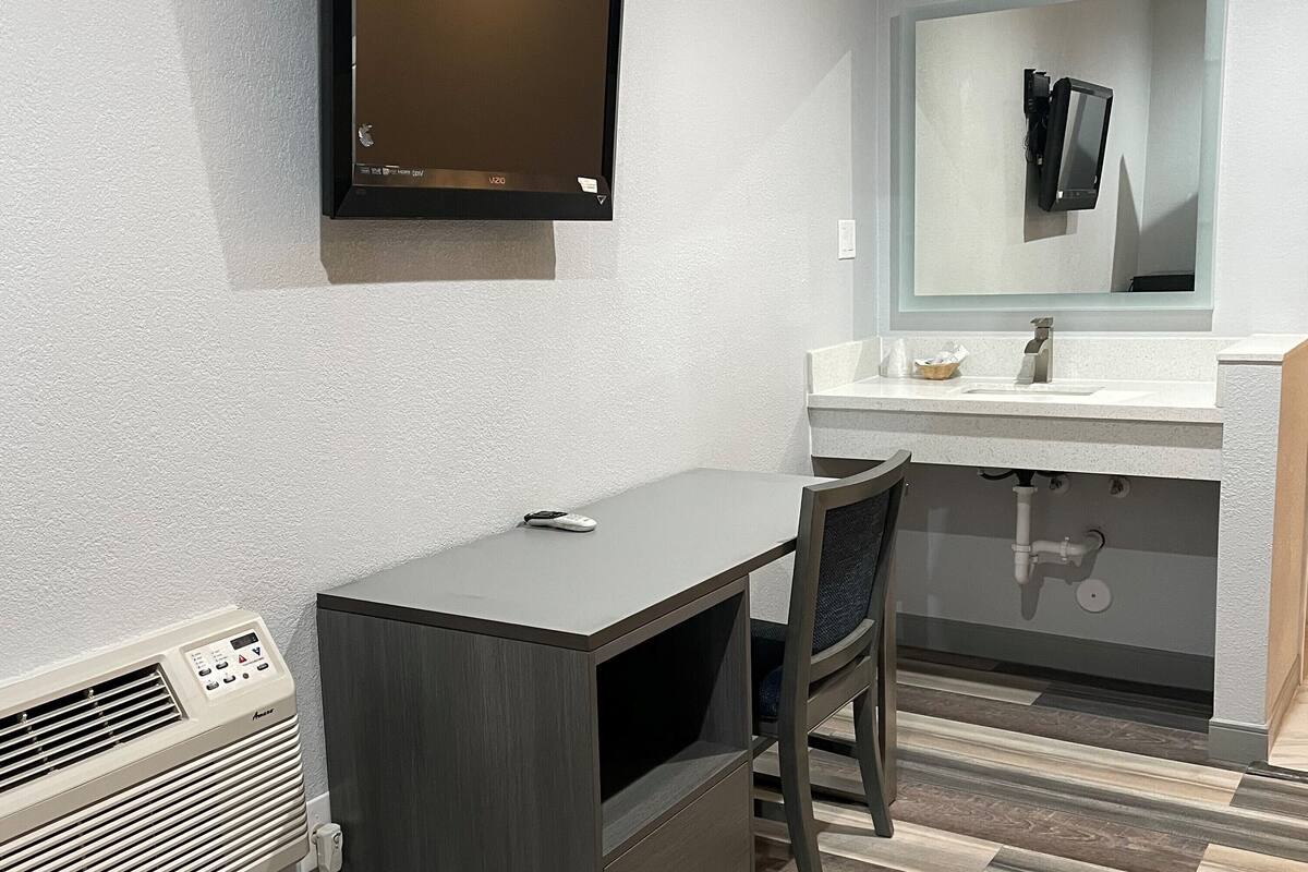 A modern bathroom with a flat-screen TV mounted on the wall, a sink with a mirror, and a desk with a chair.