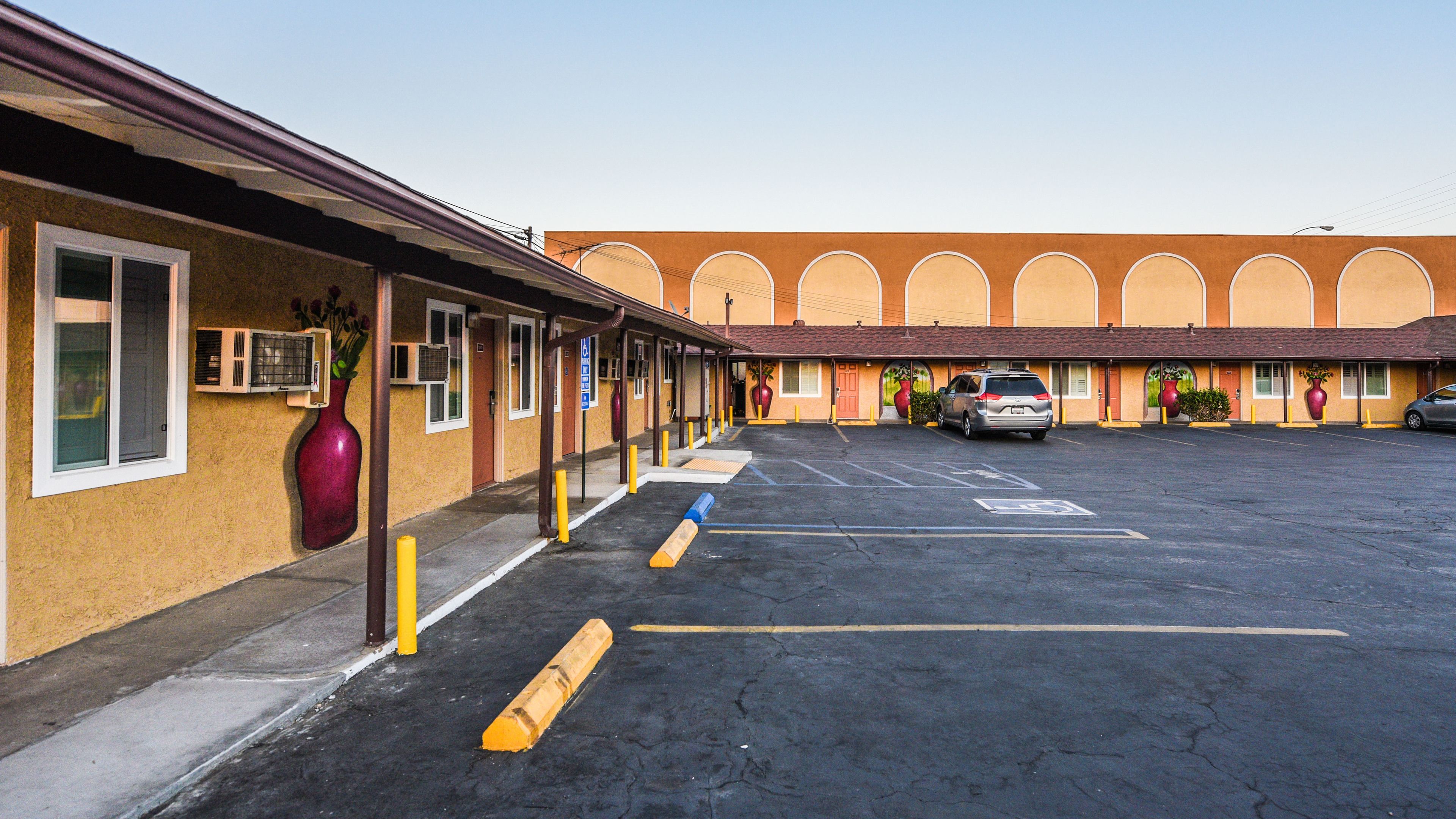 Un motel con estacionamiento, una fila de habitaciones con ventanas amplias y una fachada distintiva en arco.