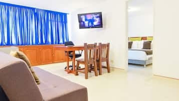 Un salon avec un canapé, une table à manger et des chaises, un téléviseur fixé au mur, et un lit dans la pièce adjacente.
