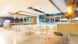 Un restaurante moderno con sillas y mesas blancas, una zona de bar y grandes ventanales con vistas a la ciudad.