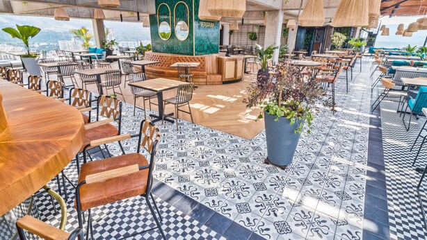 Un bar en la azotea con piso a cuadros, mesas y sillas de madera, rodeado de luces colgantes y plantas en macetas.