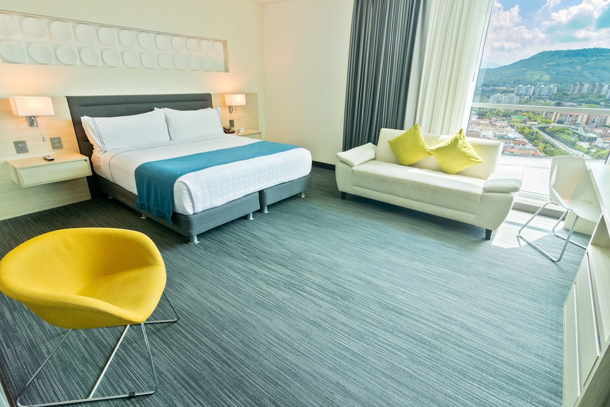 Une chambre d’hôtel avec un lit, un canapé, un fauteuil jaune et une vue sur la ville.