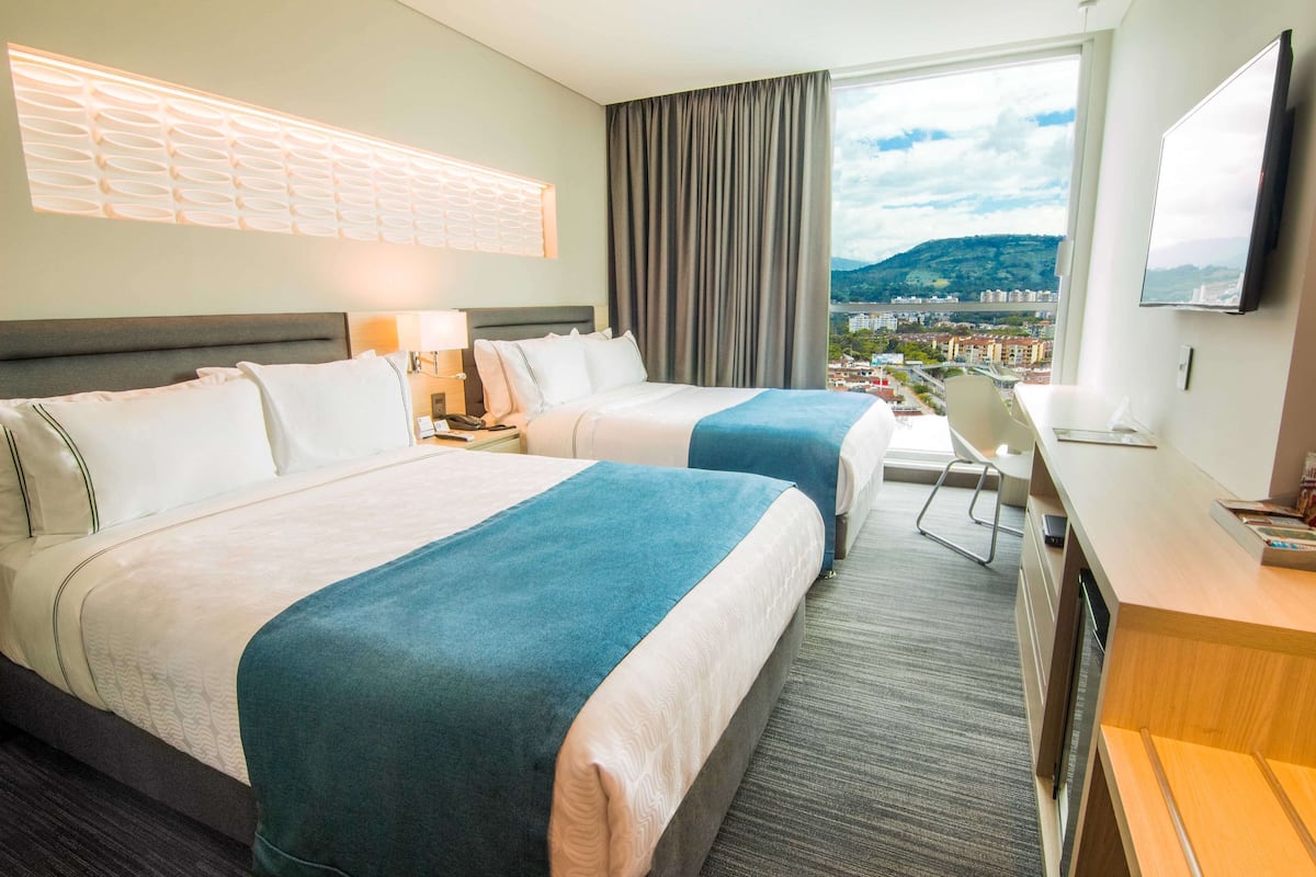 Une chambre d’hôtel avec deux lits, un bureau, une chaise et une grande fenêtre donnant sur la ville.