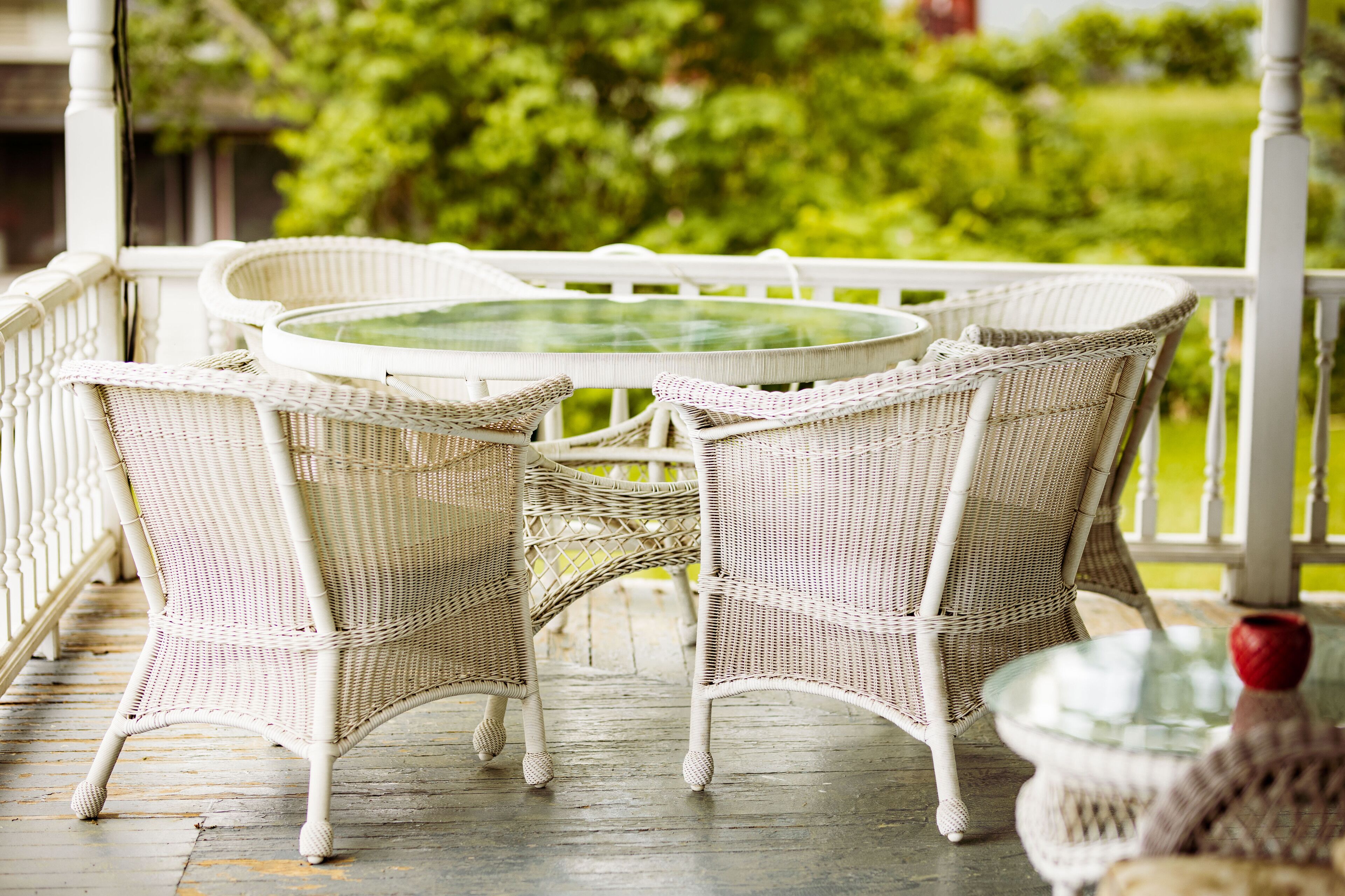 A white wicker table and chairs on a wooden porch with a glass top, surrounded by greenery.