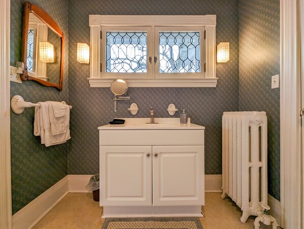 Un baño con tocador blanco, un espejo, una ventana con celosía decorativa y un radiador.