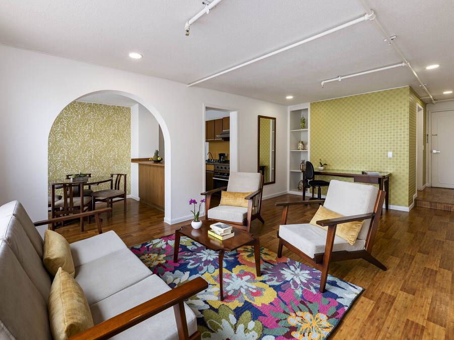 Amplia sala de estar con un sofá, sillones, una mesa de centro y un tapete colorido. Se observa una cocina al fondo con armarios de madera y un comedor con mesa y sillas.