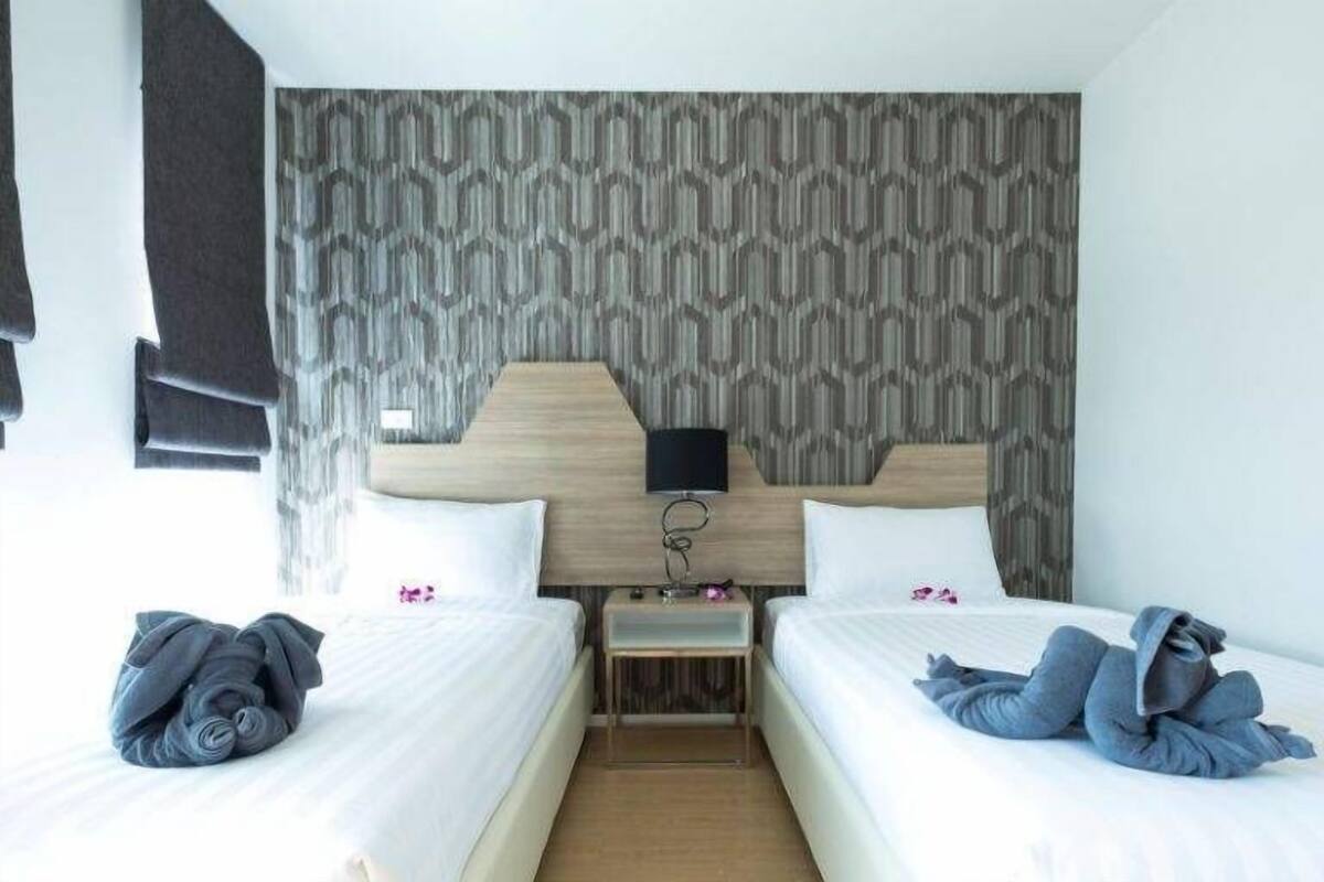 Hotellihuone, jossa on kaksi sänkyä, kuviollisella tapetilla päällystetty sängynpääty, yöpöytä lampun kanssa ja puulattia.
