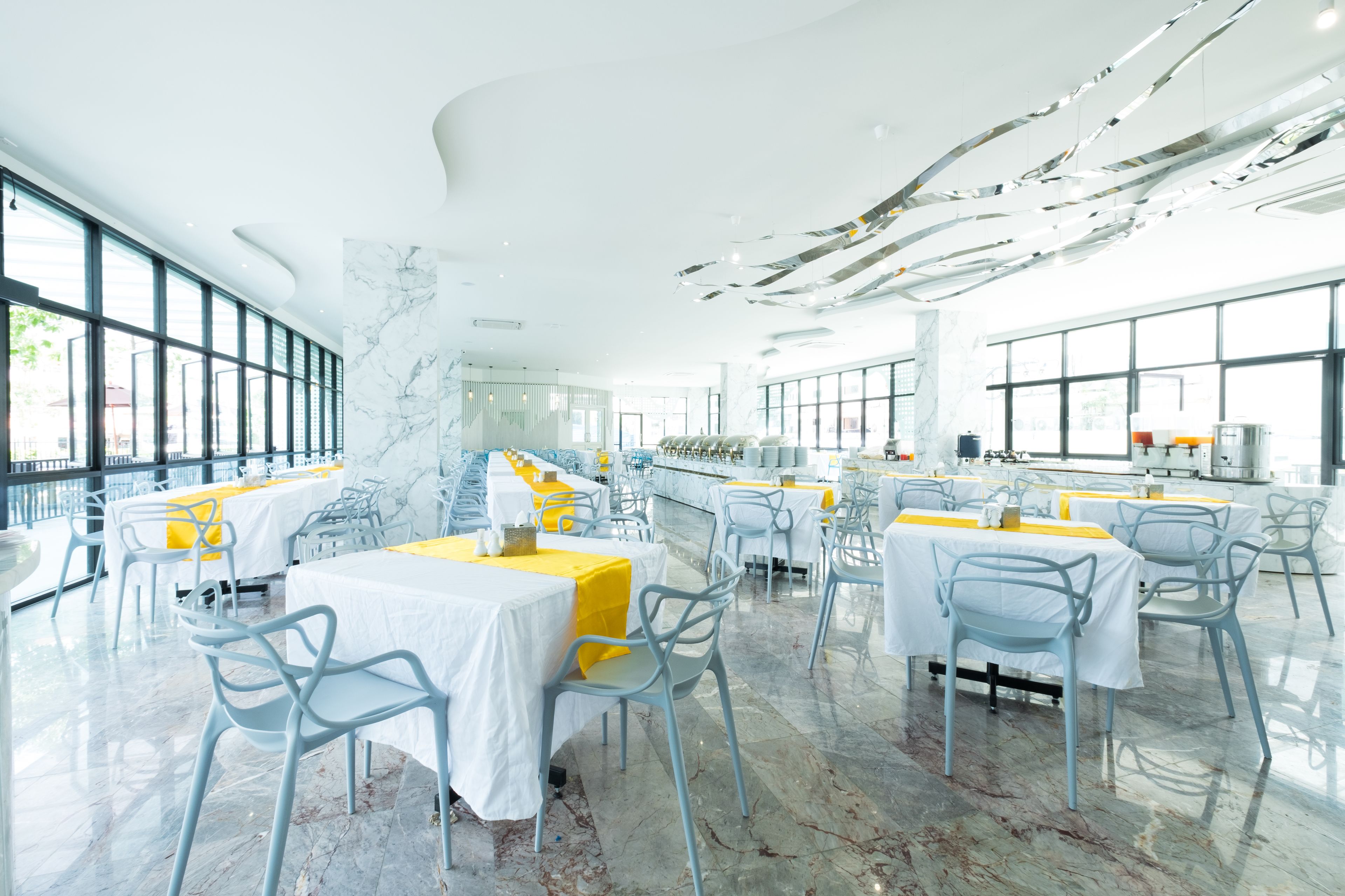 A spacious, modern dining area with marble floors and large windows, featuring tables with yellow tablecloths and light blue chairs.