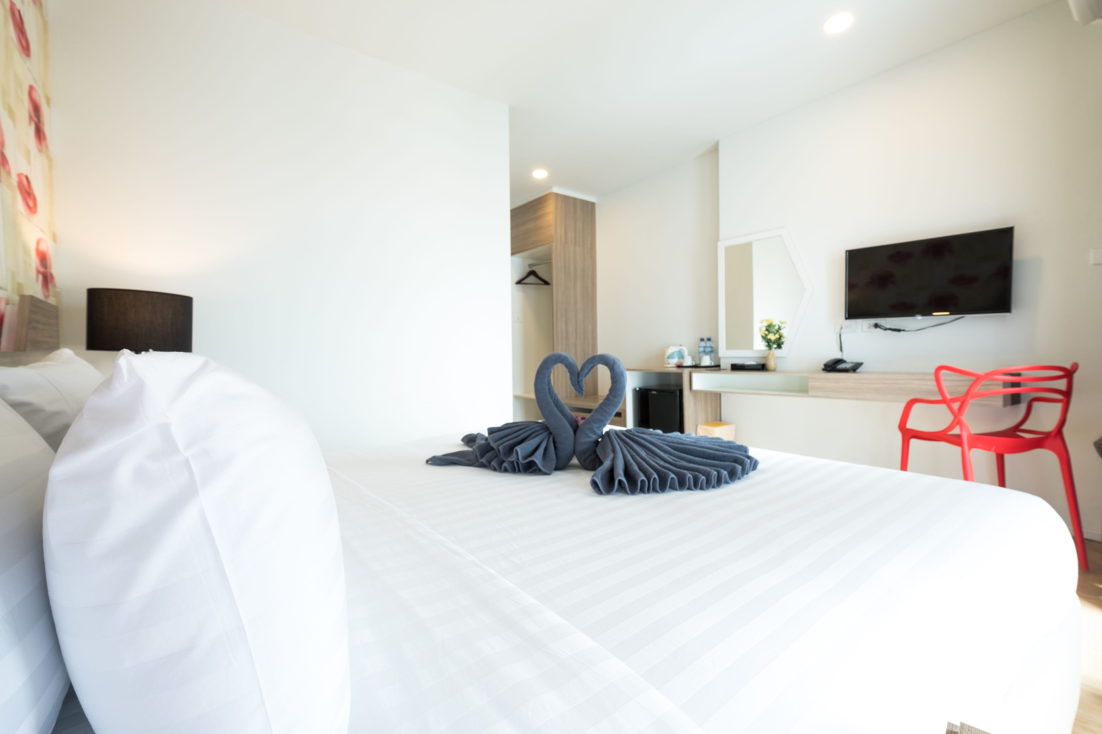 A hotel room with a neatly made bed, a flat-screen TV mounted on the wall, a desk with a chair, and a red chair.