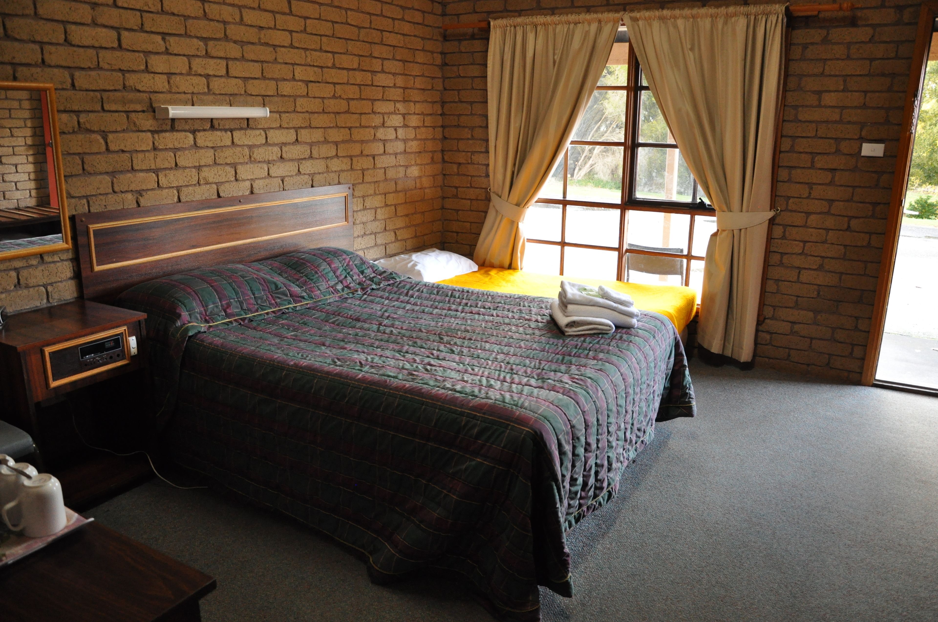 Ein Zimmer mit Backsteinwand, einem Bett mit Steppdecke, einem Nachttisch mit Radio und einem Fenster mit Vorhängen.
