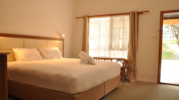 A neatly made bed with white linens and a rolled towel, a bedside table with a lamp, and a window with sheer curtains. - Miner's Retreat Motel (Ballarat East)