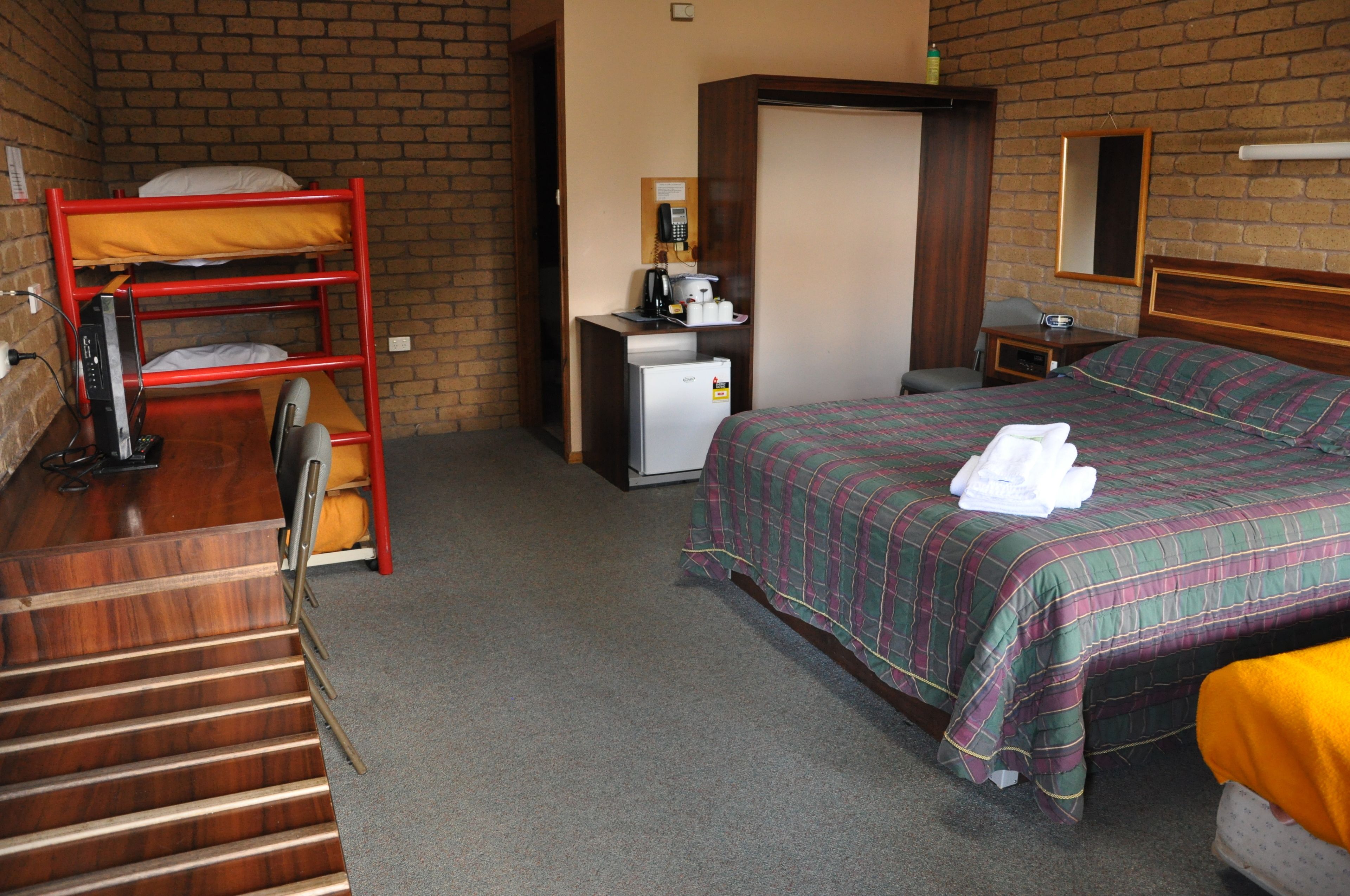 A hotel room with a bunk bed, a small desk with a refrigerator, a bedside table with a lamp, and a large bed with a plaid bedspread.