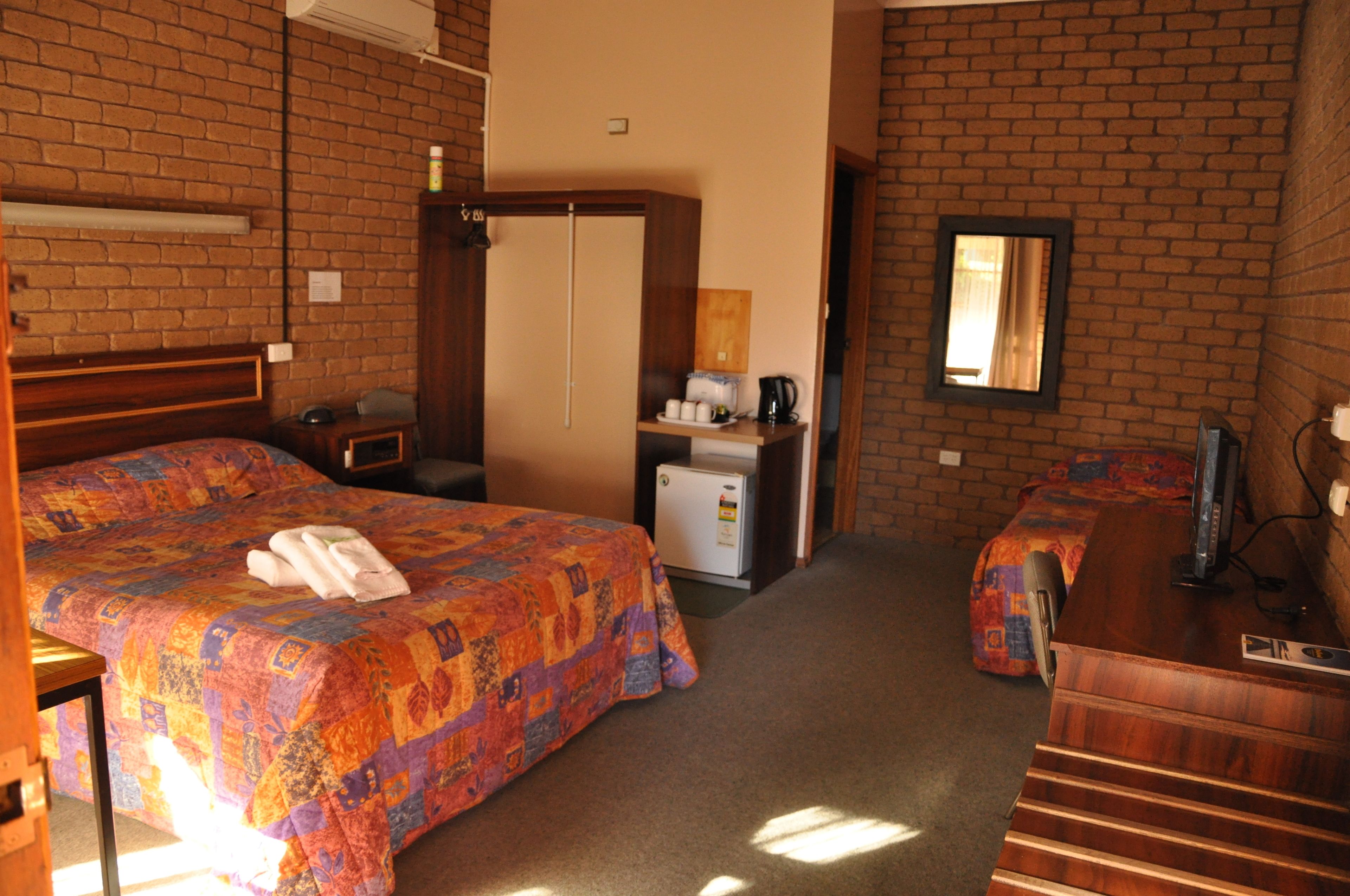 A hotel room with a brick wall, a bed with a colorful quilt, a desk with a computer, and a small refrigerator.