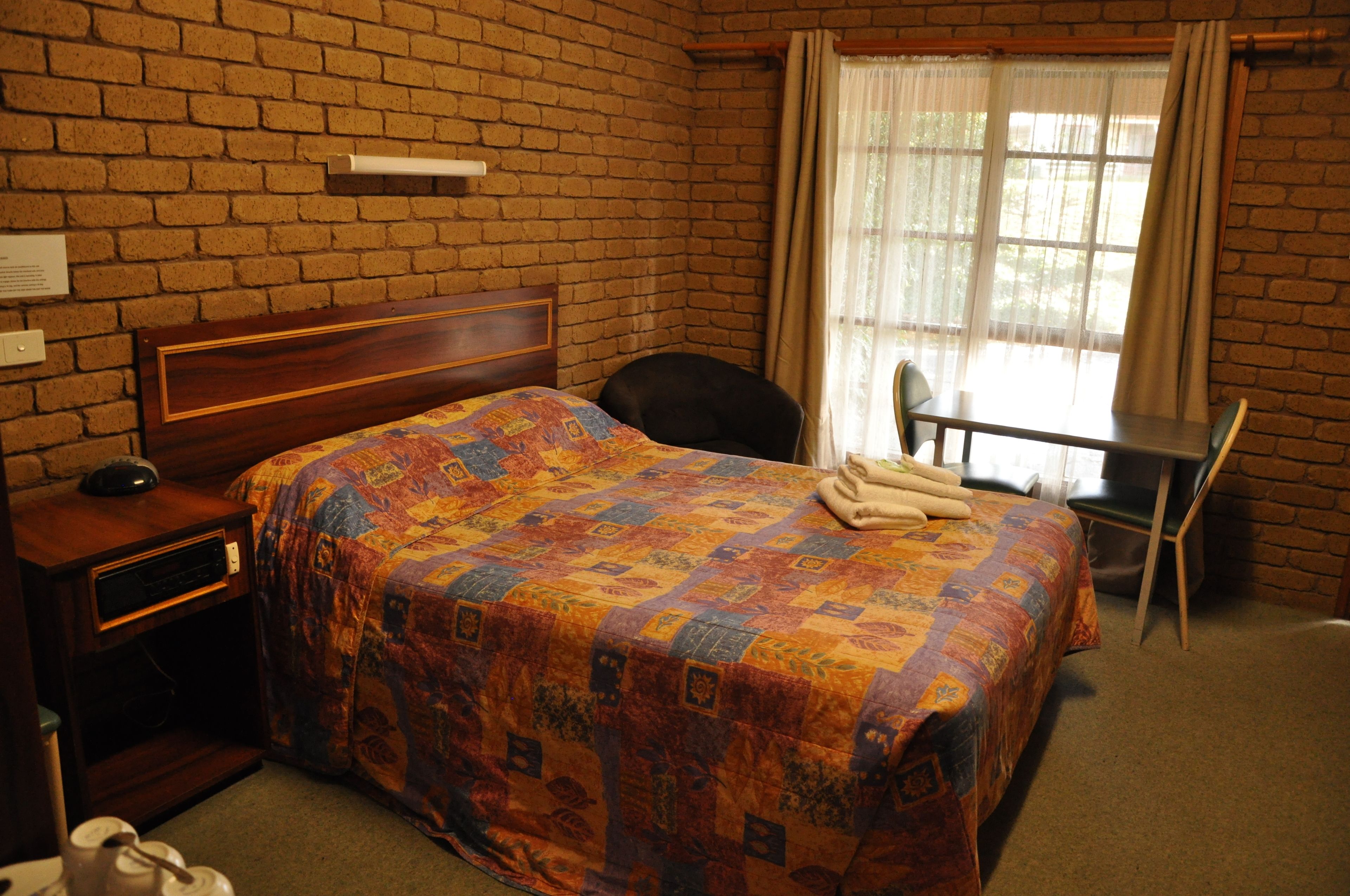 A hotel room with a brick wall, a wooden bed with a colorful quilt, a desk with a chair, and a window with curtains.