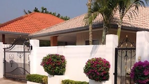 A house with a white exterior, a red-tiled roof, and a gated entrance. There are three flower planters with pink flowers and a palm tree in the background. - Baansiesom Private Pool Villa (Pranburi)