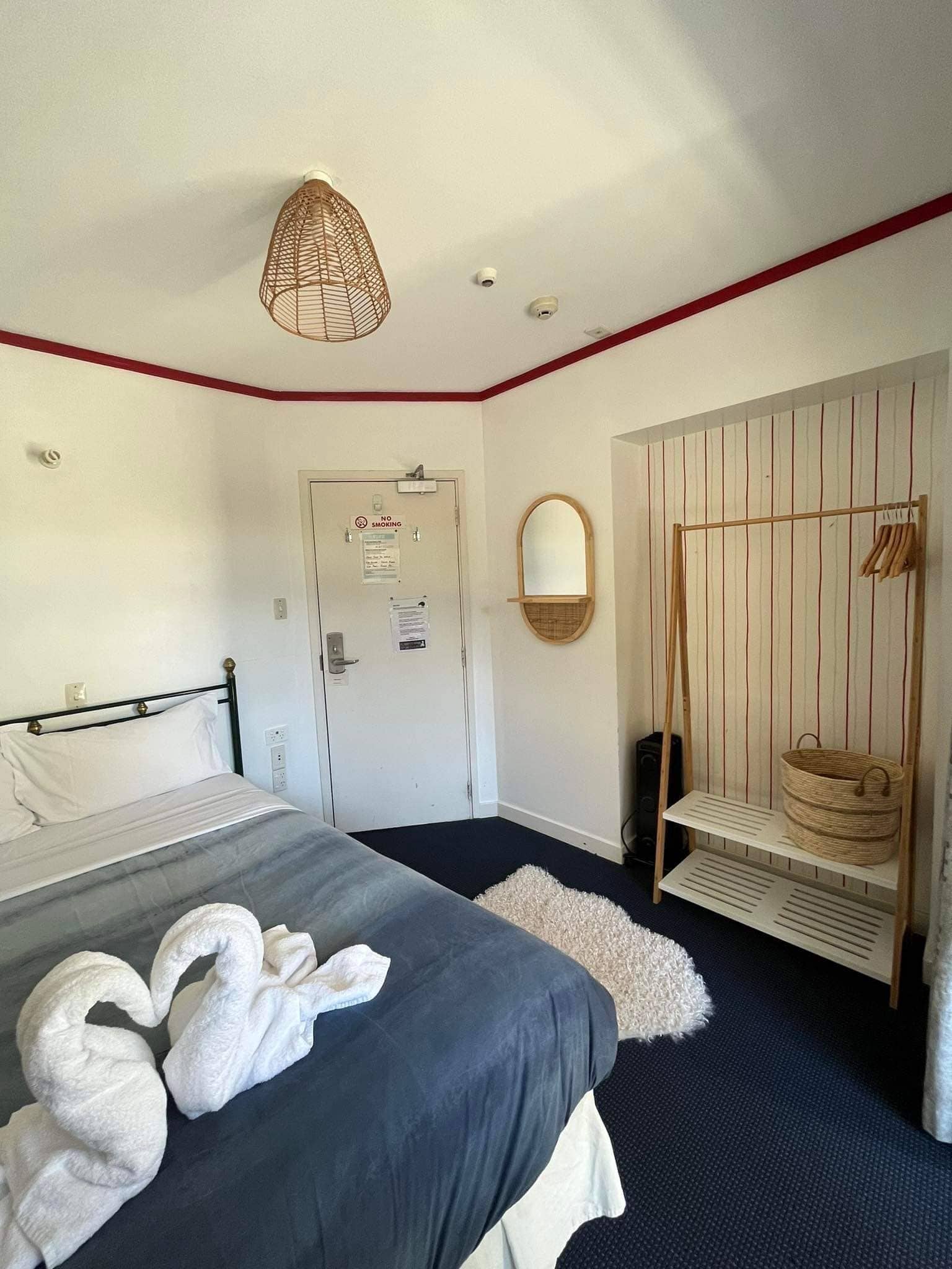 A hotel room with a bed, a door with a 'No Smoking' sign, a mirror, a shelf with baskets, and a hanging light fixture.