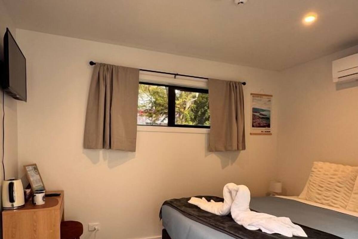A bedroom with a bed, a desk with a chair, a window with curtains, and a television on the wall.
