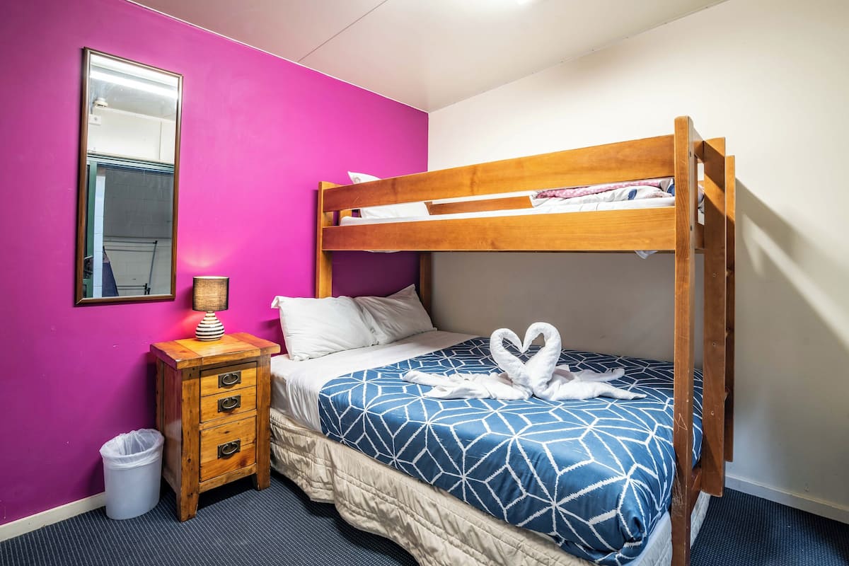 A bunk bed with a geometric-patterned blue and white bedspread, a wooden nightstand with a lamp, a mirror, and a trash bin.