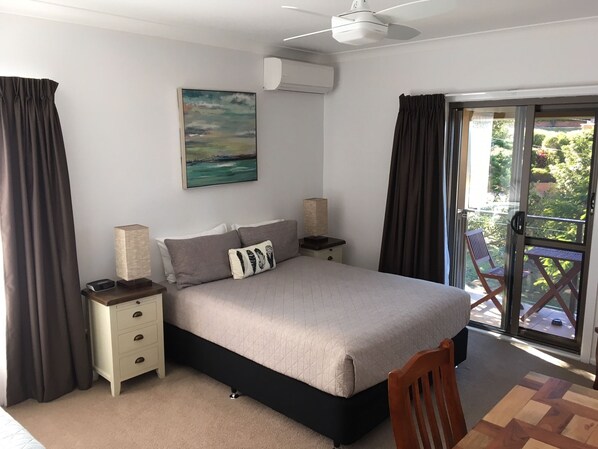 A bedroom with a bed, bedside tables, a painting, an air conditioner, and a view of the outdoors.