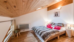 Un dormitorio con techo de madera, una cama con una manta de piel, almohadas rojas, un taburete pequeño, un radiador y un cuadro en la pared.