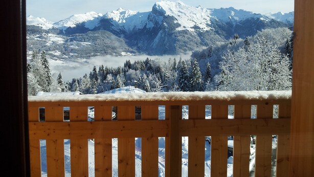 Vista desde un balcón de madera con montañas y árboles cubiertos de nieve al fondo.