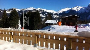 Una valla de madera con suelo cubierto de nieve, una cabaña de madera con techo nevado y montañas nevadas al fondo.