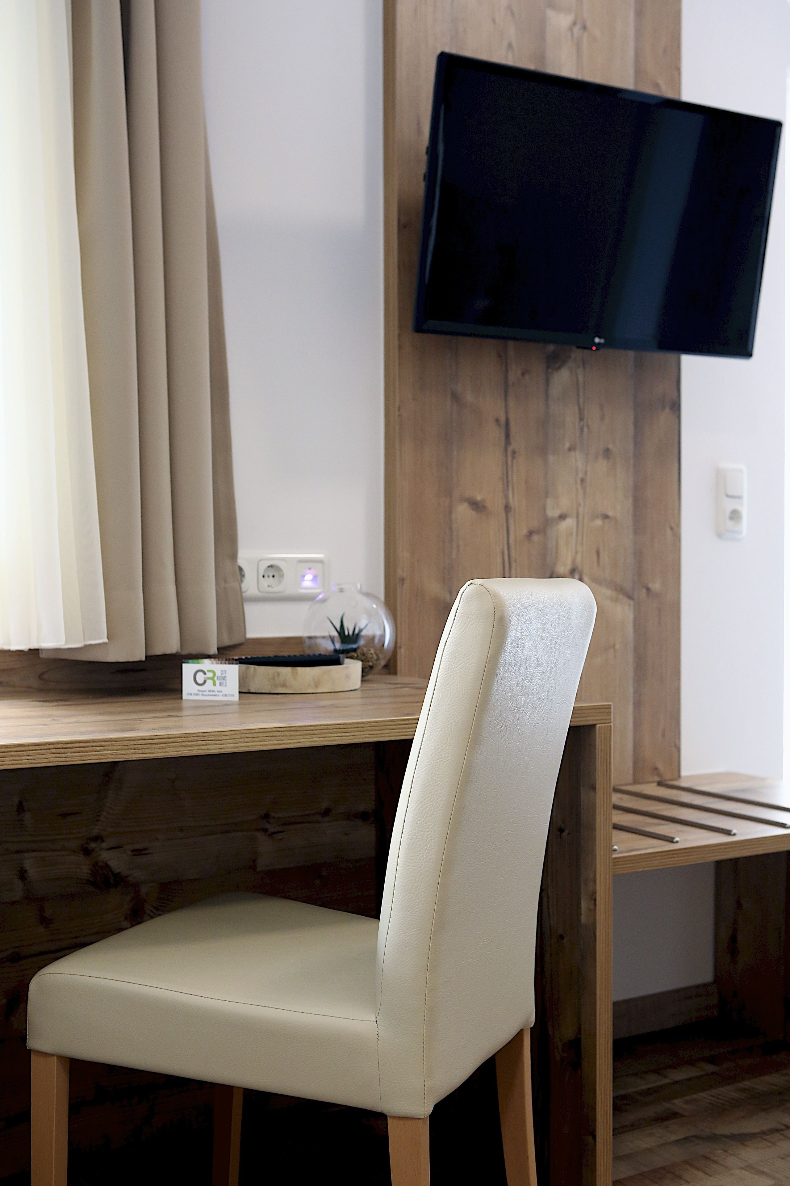 Ein modernes Zimmer mit einem Holzbüro, einem weißen Stuhl, einem an der Wand montierten Flachbildfernseher und einer kleinen Zimmerpflanze auf dem Schreibtisch.