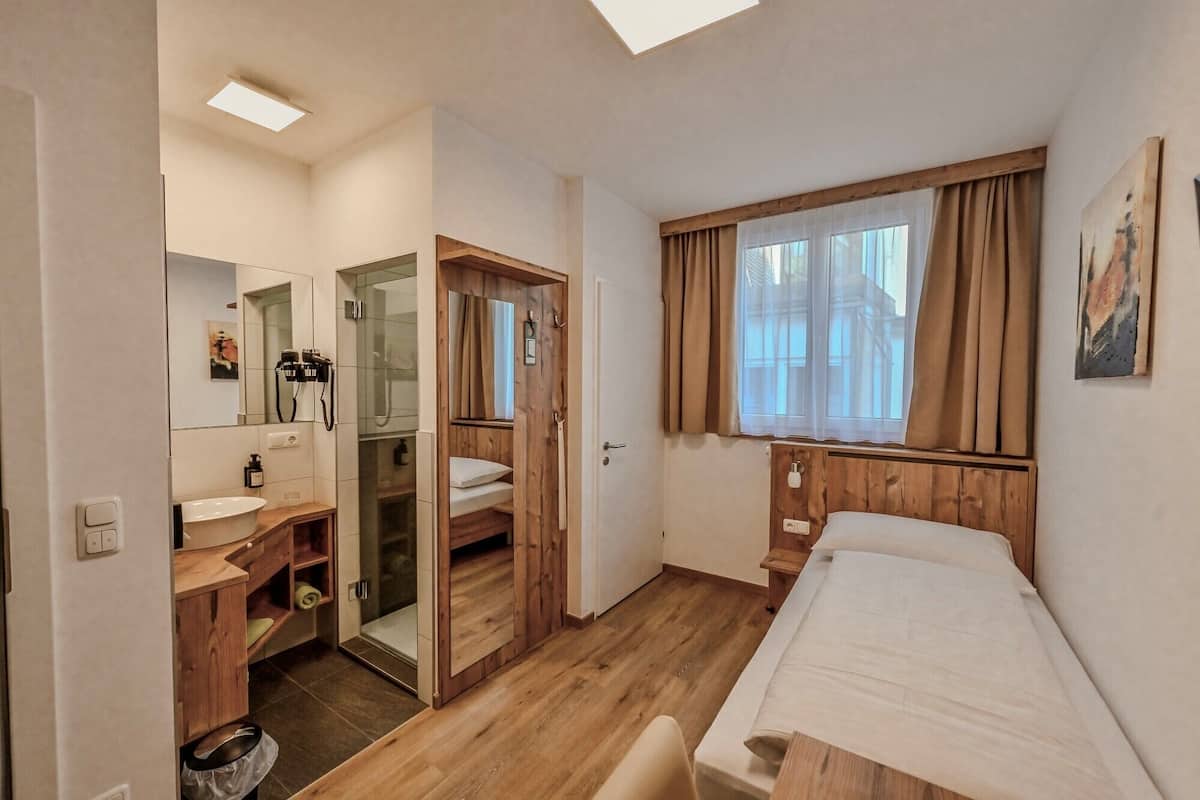 Une chambre à coucher avec un lit, une fenêtre avec des rideaux, une armoire en bois, un miroir et une salle de bain avec un lavabo et une douche.
