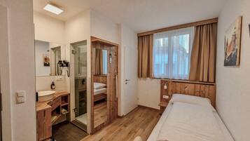 Une chambre à coucher avec un lit, une fenêtre avec des rideaux, une armoire en bois, un miroir et une salle de bain avec un lavabo et une douche.