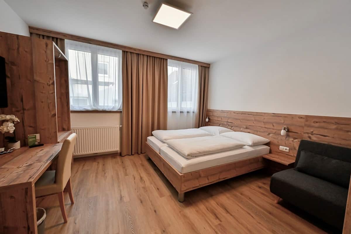 Une chambre d’hôtel avec un plancher en bois, un lit avec du linge de maison blanc, un bureau avec une chaise, une petite table avec un vase de fleurs et une fenêtre avec des rideaux.
