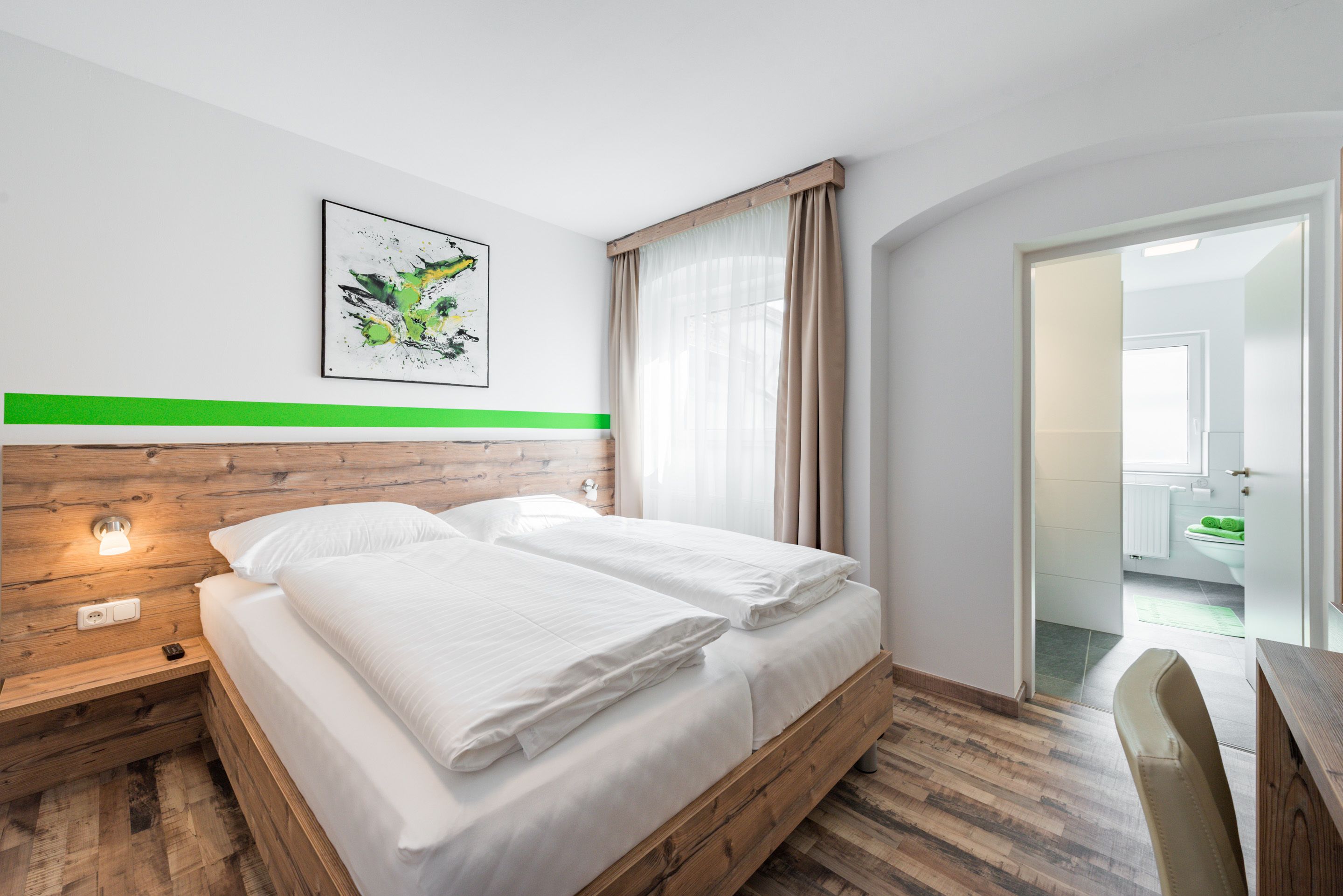 Ein modernes Hotelzimmer mit einem hölzernen Kopfteil, einem grünen Akzentstreifen, einem gerahmten abstrakten Gemälde und Blick ins Badezimmer durch eine geöffnete Tür.