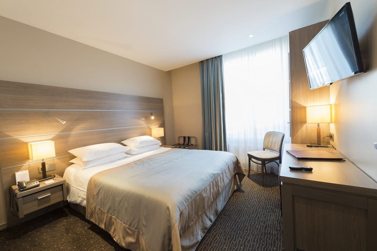 Une chambre d’hôtel comprenant un lit, des tables de chevet, un bureau, une chaise, une télévision et une fenêtre avec des rideaux.