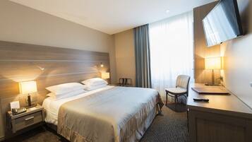 A hotel room with a bed, bedside tables, a desk, a chair, a TV, and a window with curtains.
