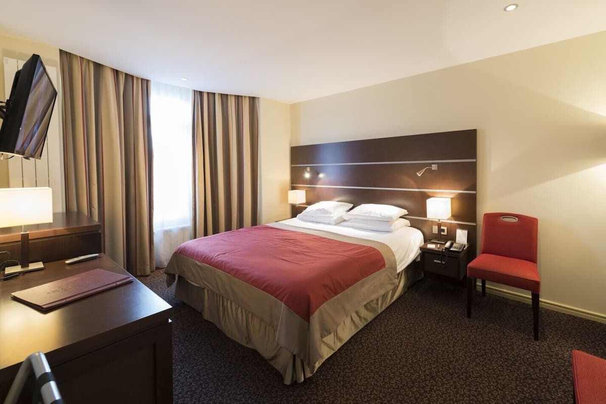 Une chambre d’hôtel comprenant un lit, une table de chevet, une chaise, un bureau, une télévision et des rideaux.