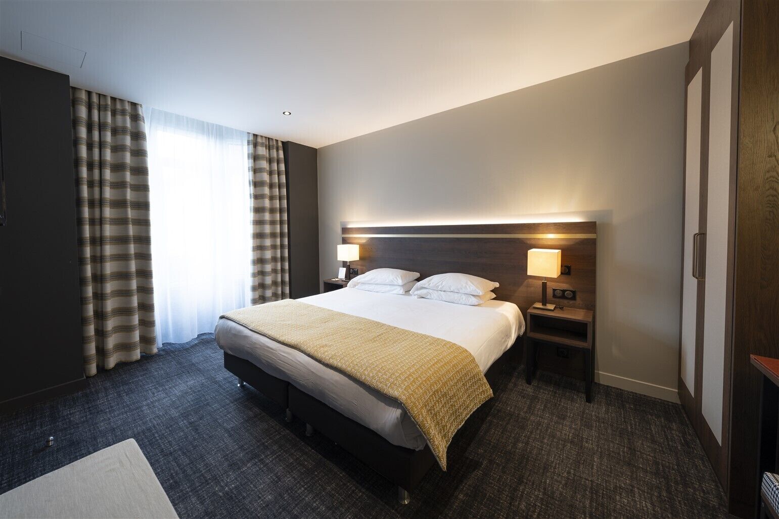 A hotel room with a large bed, two bedside tables with lamps, a dark-colored wardrobe, and a window with curtains.