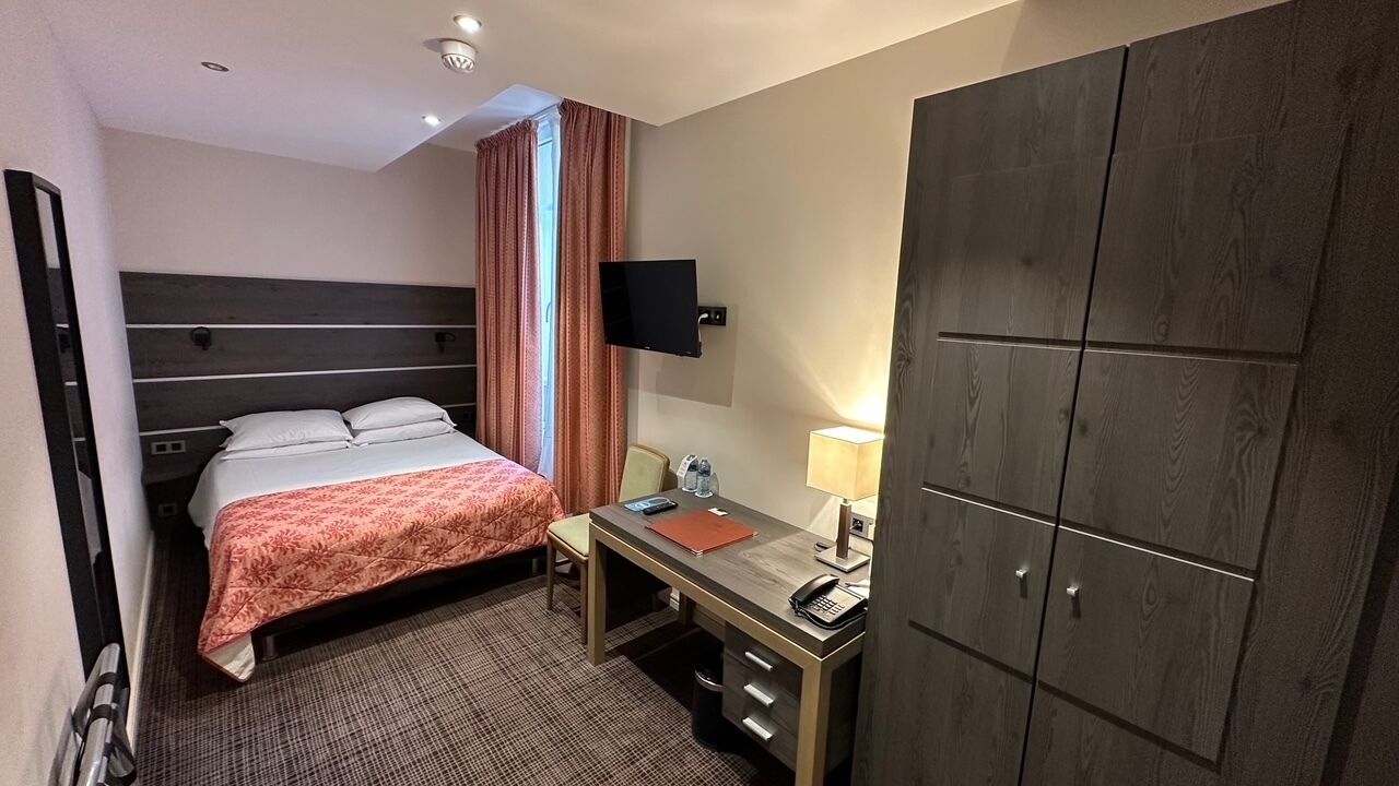 Habitación de hotel con cama, escritorio, silla, armario y televisor montado en la pared.