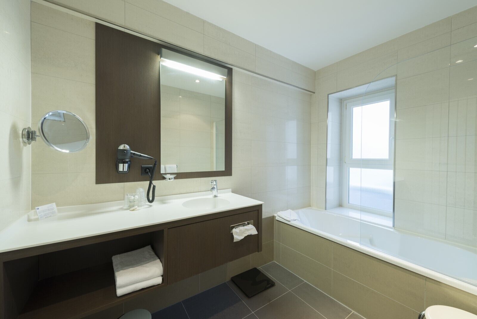 A modern bathroom with a large mirror, a sink, a vanity, a bathtub, and a window.