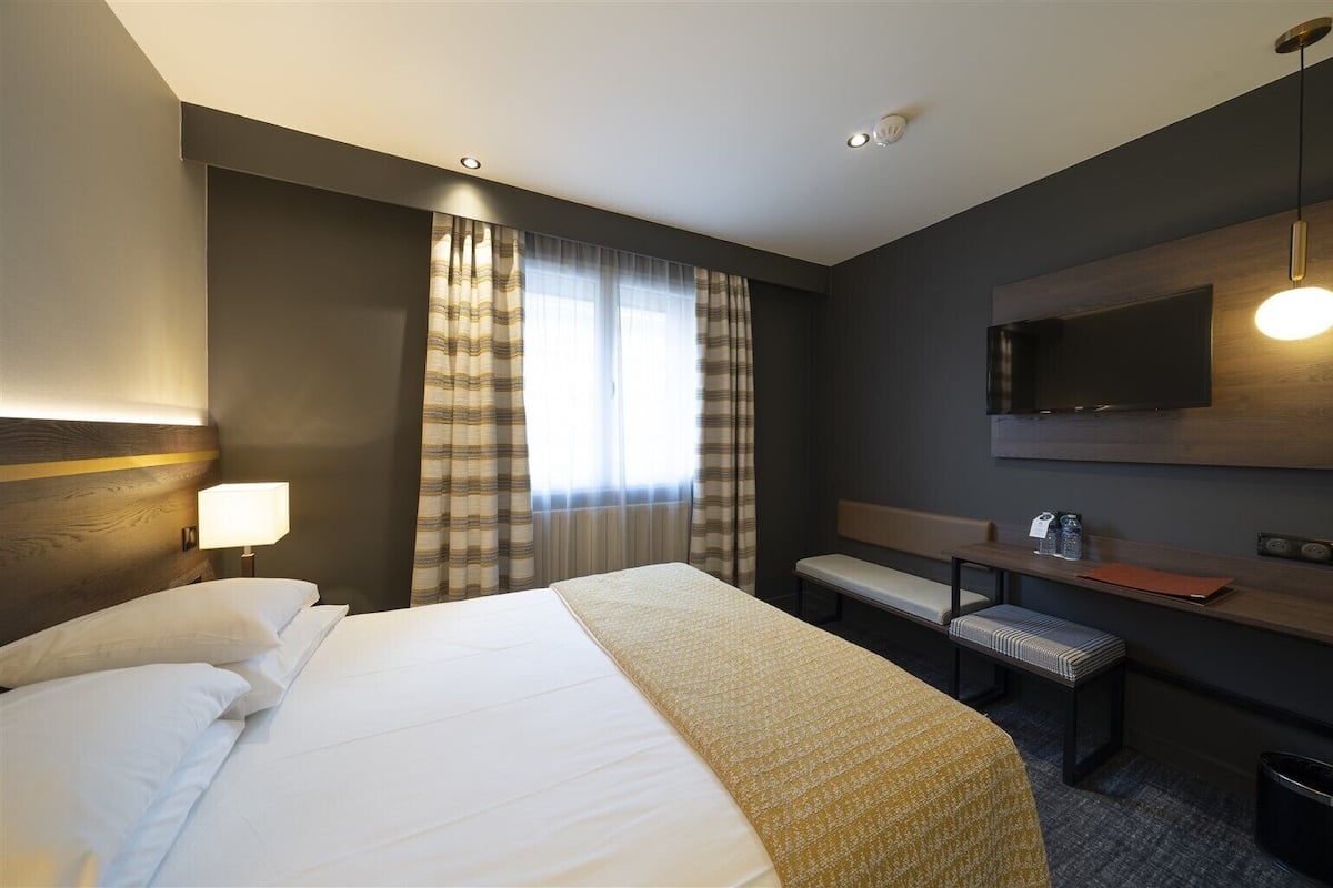 Une chambre d’hôtel avec un grand lit, un bureau avec une chaise, une télévision et une fenêtre avec des rideaux.