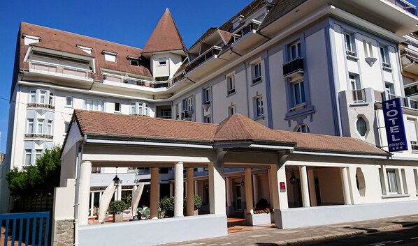 Ein Hotel im klassischen europäischen Stil mit einer markanten Eingangspforte, einer überdachten Terrasse, mehreren Balkonen und blauem Himmel im Hintergrund.