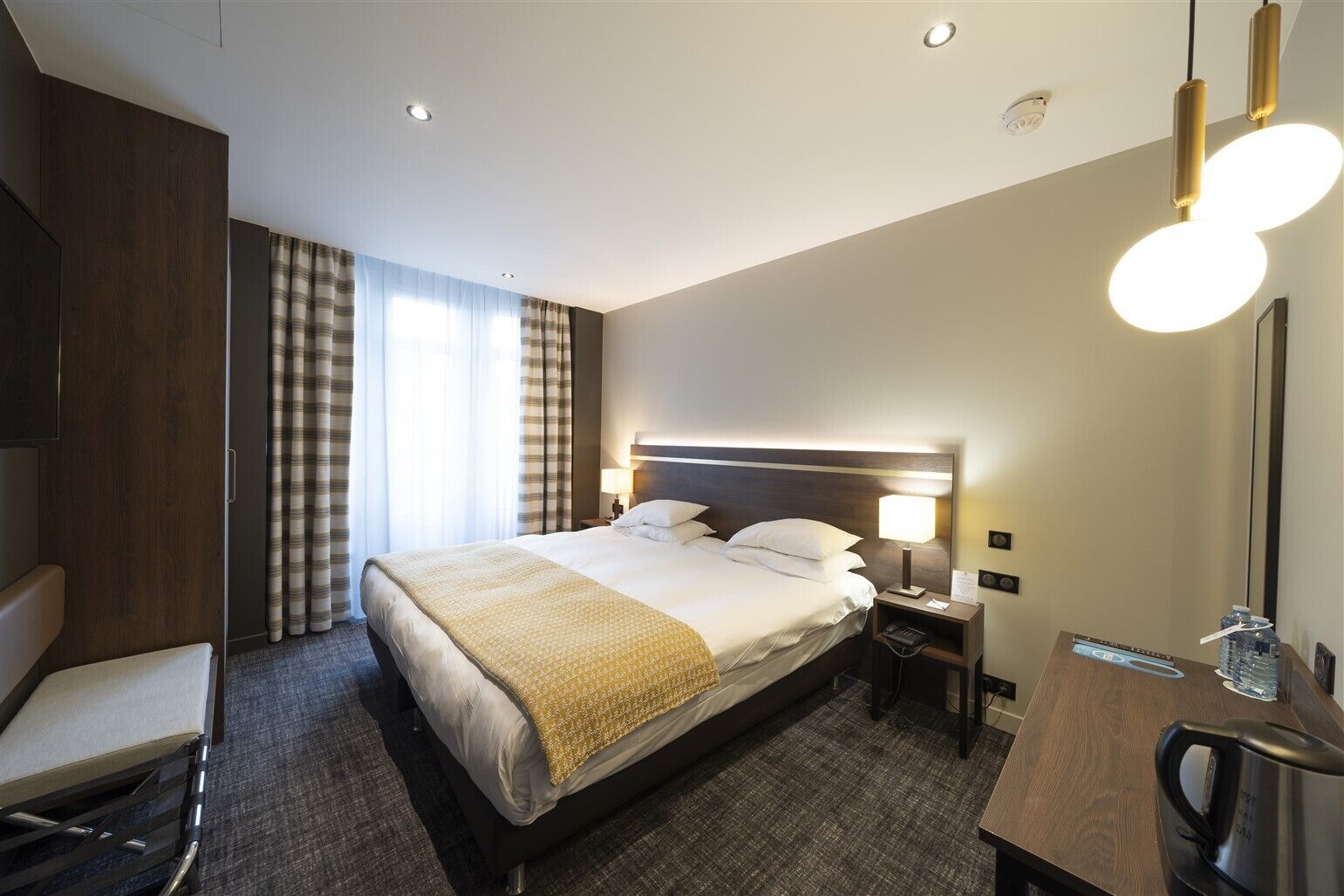 A hotel room with a large bed, bedside tables, a desk with a chair, and a modern chandelier.