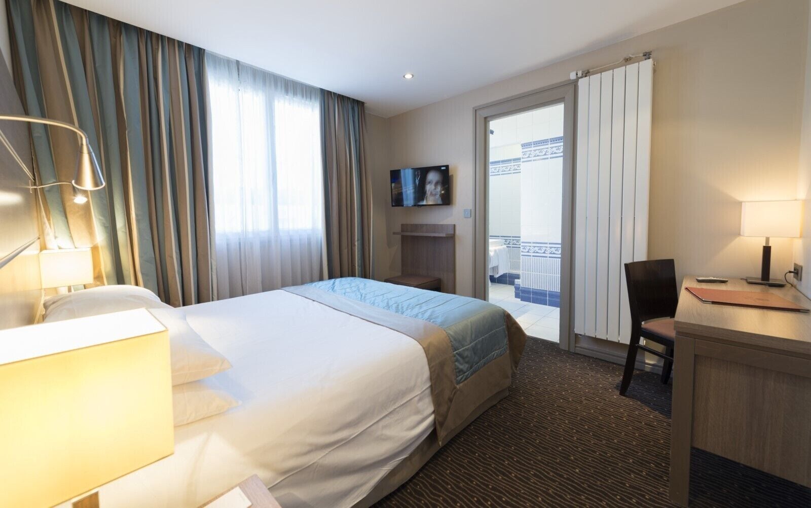 Ett hotellrum med en säng, ett skrivbord, en stol, en TV och en balkong med utsikt.