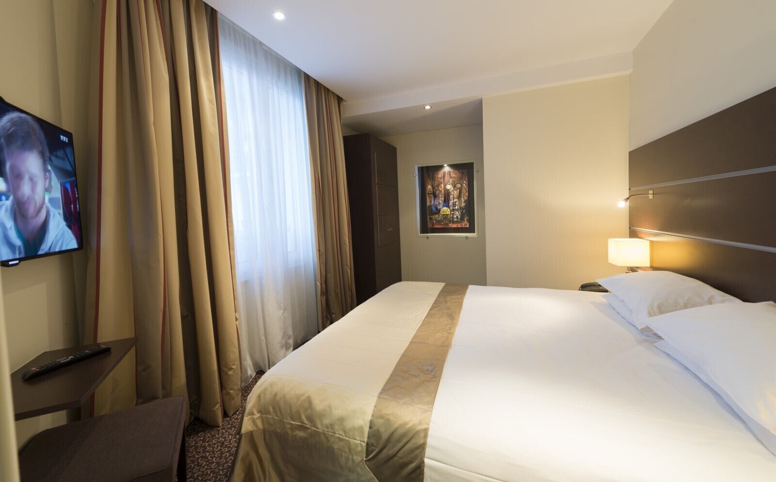 Une chambre d’hôtel avec un grand lit, un téléviseur fixé au mur, un bureau avec une chaise et un tableau accroché au mur.