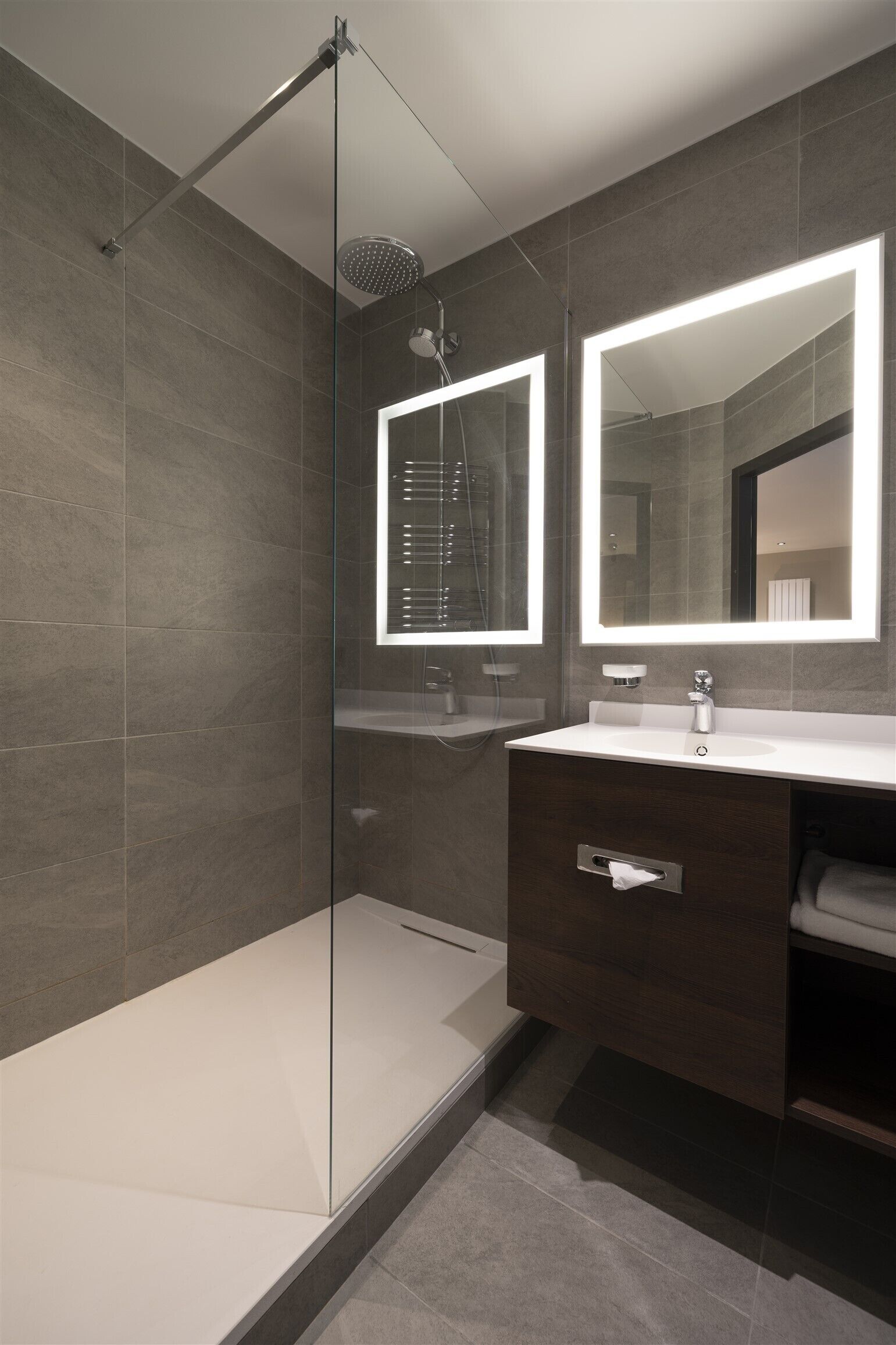 Un baño moderno con ducha envuelta en vidrio, un espejo grande y un tocador de madera oscura.