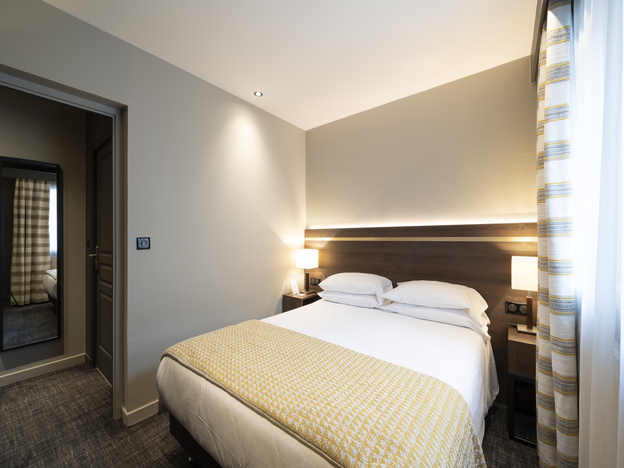 A hotel room with a bed, bedside lamps, a door, and a curtain.