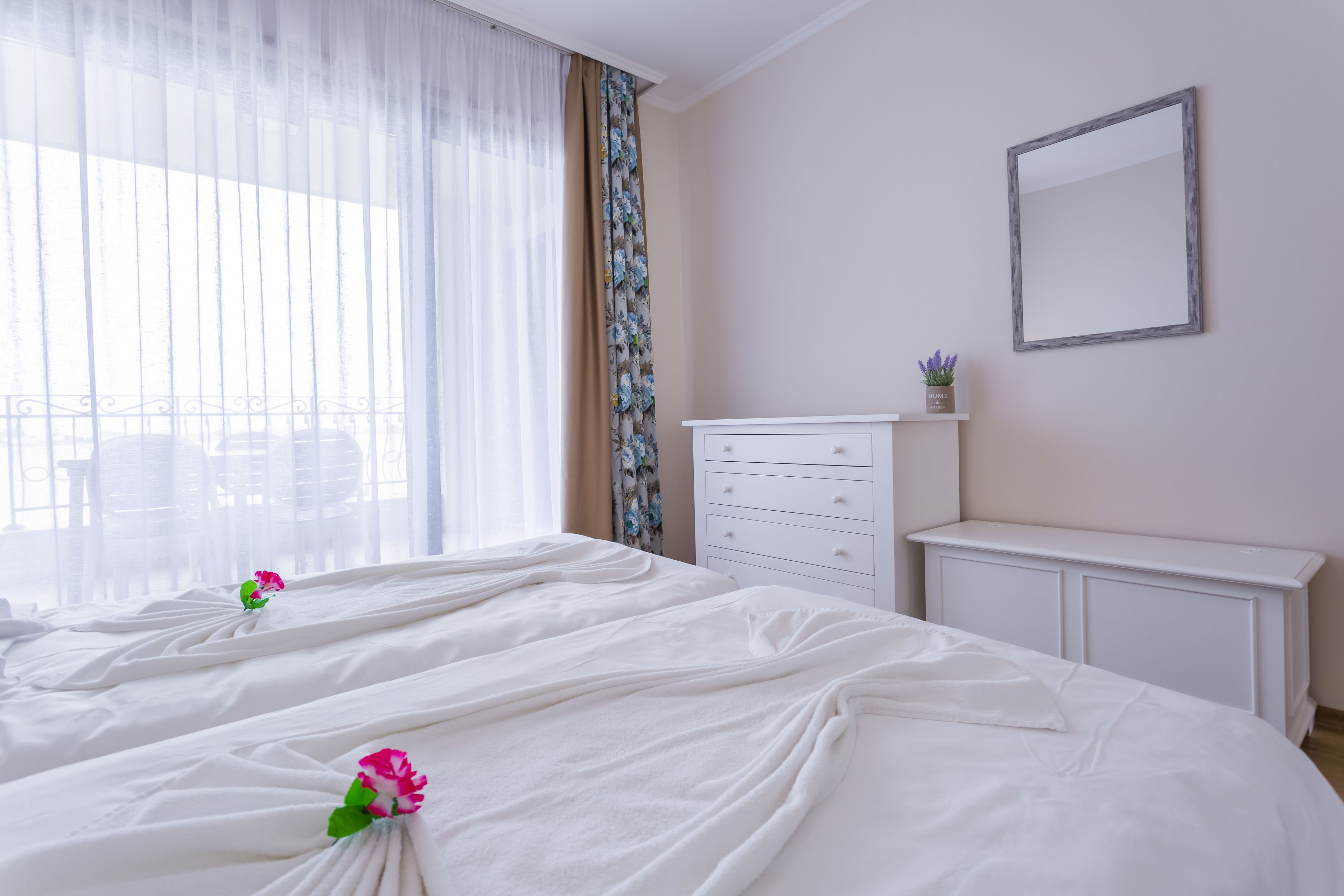 A hotel room with a large bed, a dresser, a mirror, and a window with sheer curtains.