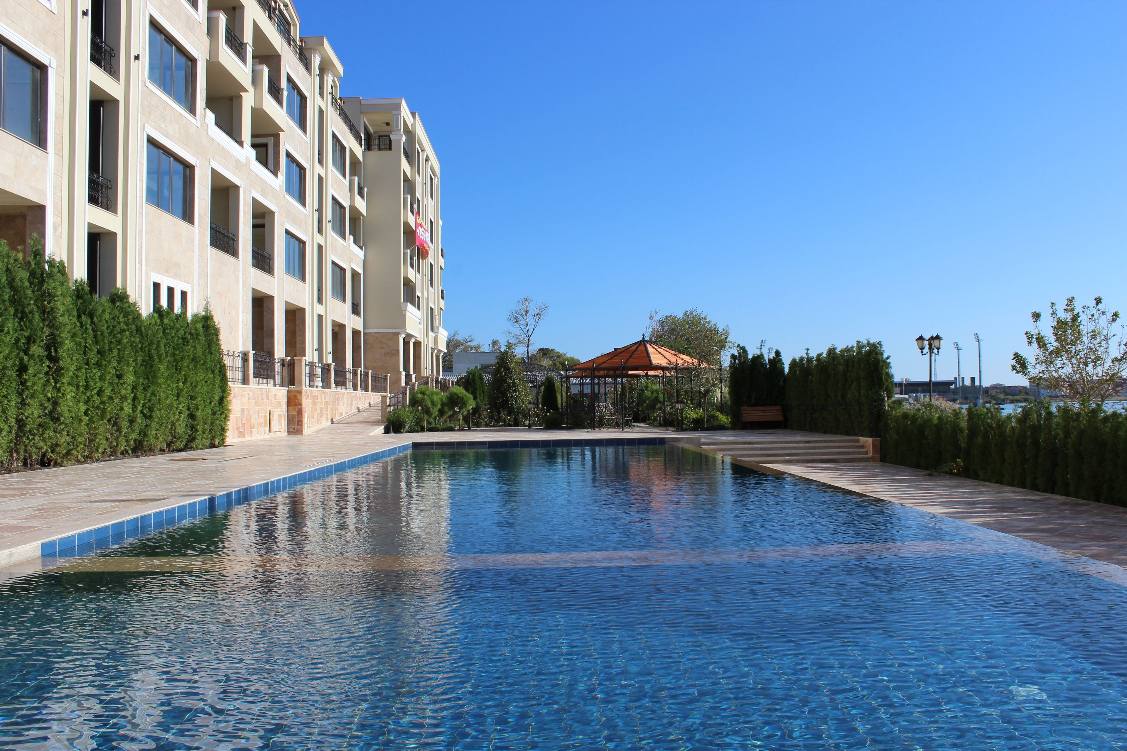 A swimming pool with a clear blue water surface, surrounded by a landscaped area with neatly trimmed hedges and a building with multiple balconies.