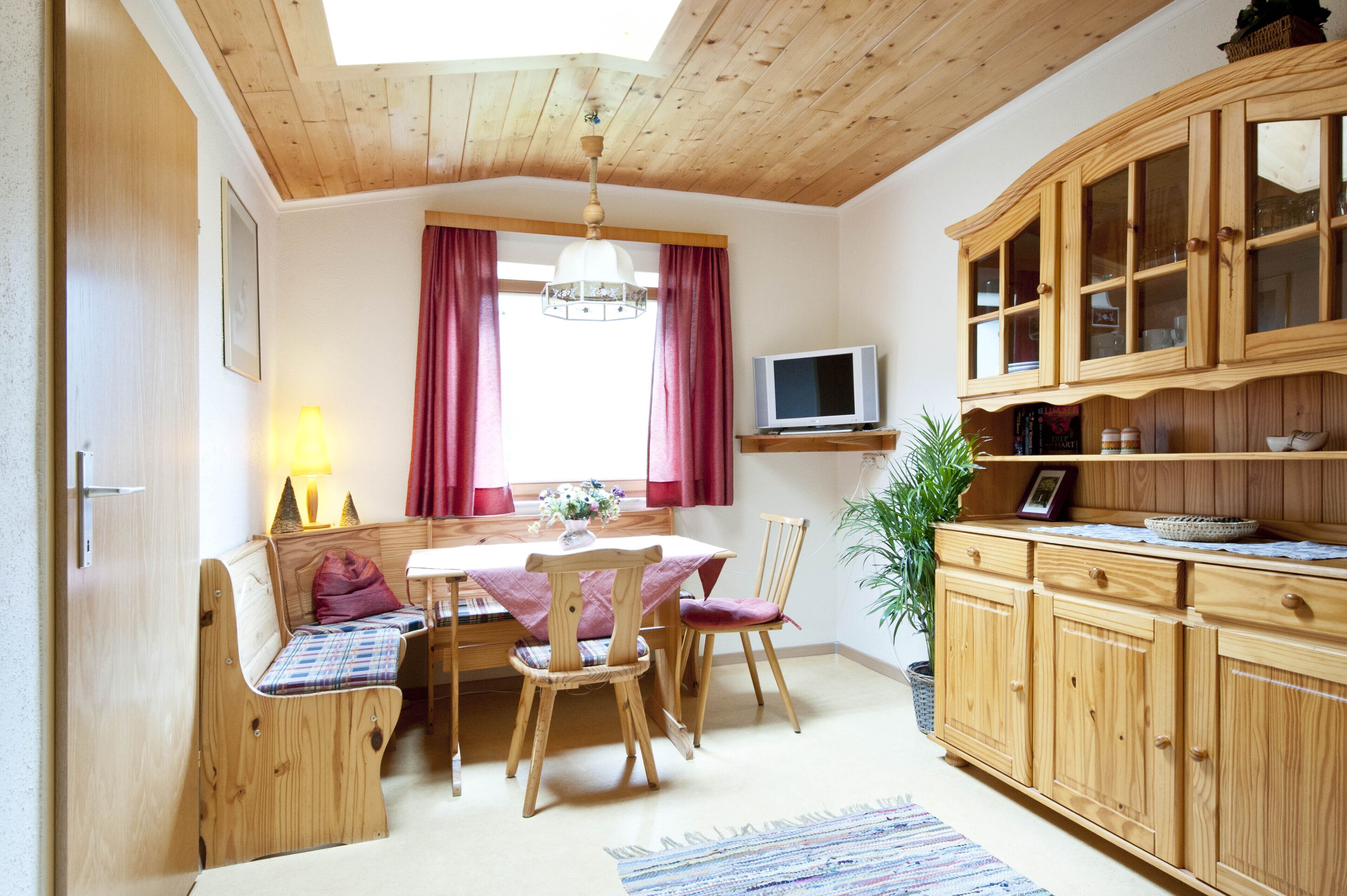 Een gezellige kamer met een houten plafond, een eettafel met stoelen, een televisie op een plank en een kast met glazen deuren.
