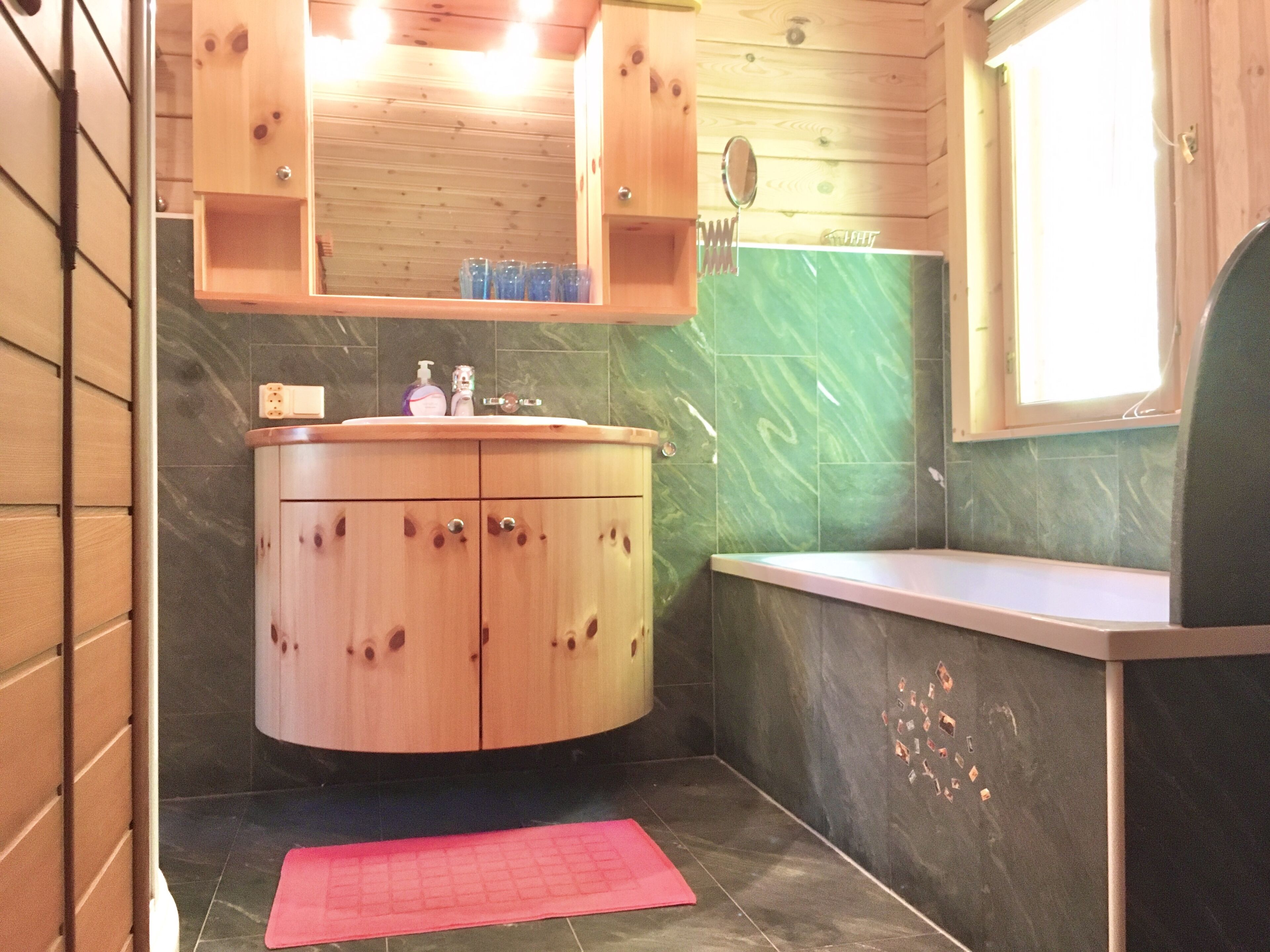 A bathroom with wooden cabinets, a marble-tiled wall, a sink, and a window.